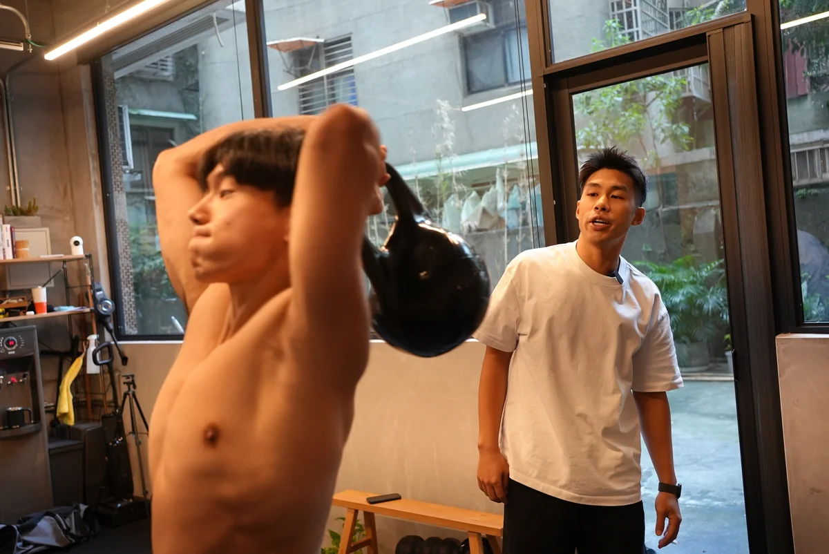 窗邊自然光下,學員做壺鈴過頭動作,Taka 在一旁看著